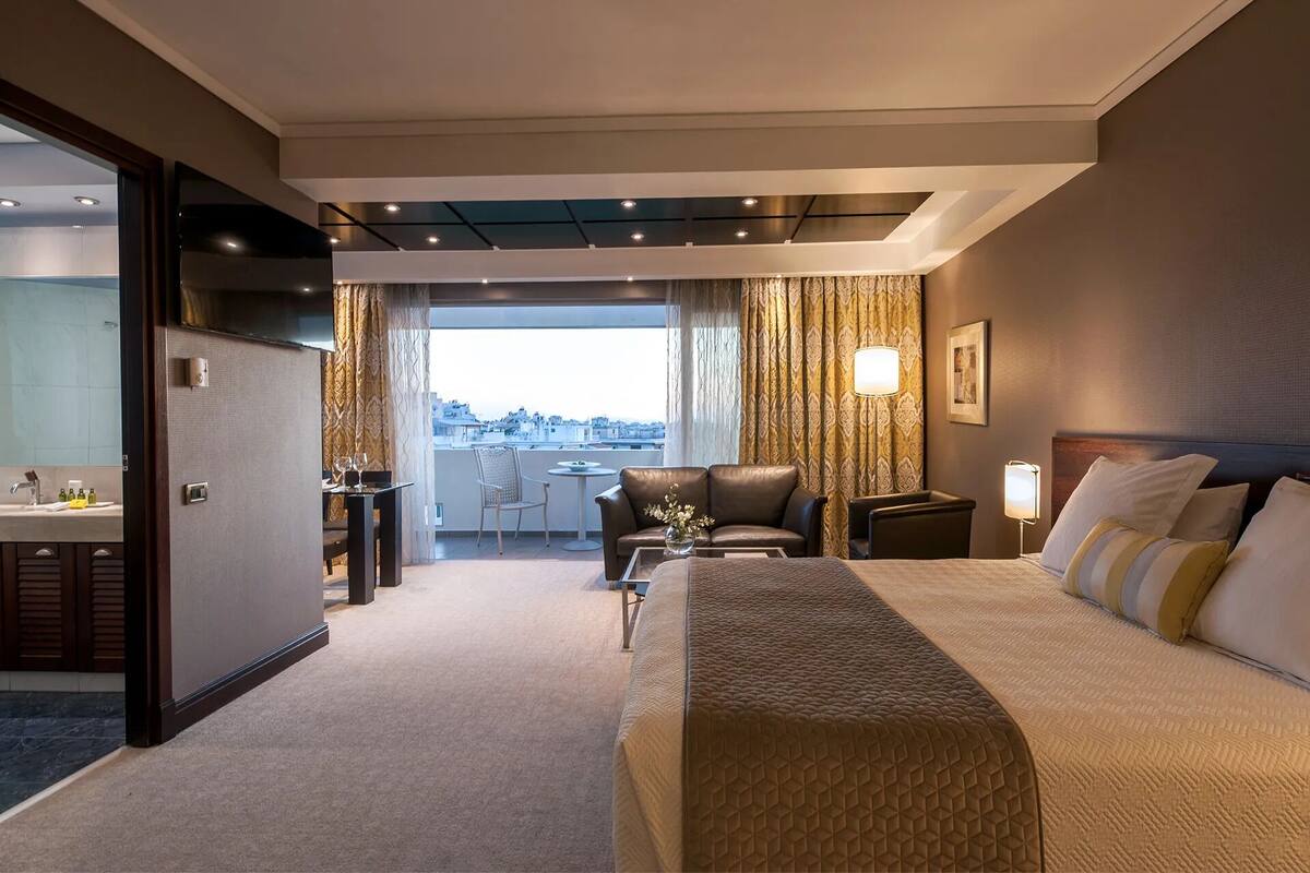 A hotel room with a bed, a desk, a chair, a sofa, and a view of the city.
