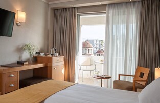 Quarto de hotel com cama, escrivaninha com luminária, cadeira e vista para prédios pela janela.