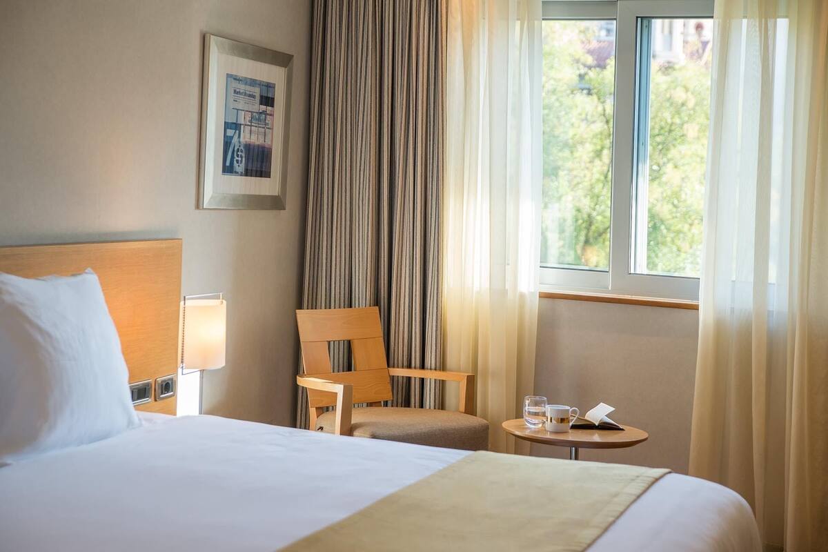 A hotel room with a bed, a chair, a small table with a book and a glass, and a window with curtains.
