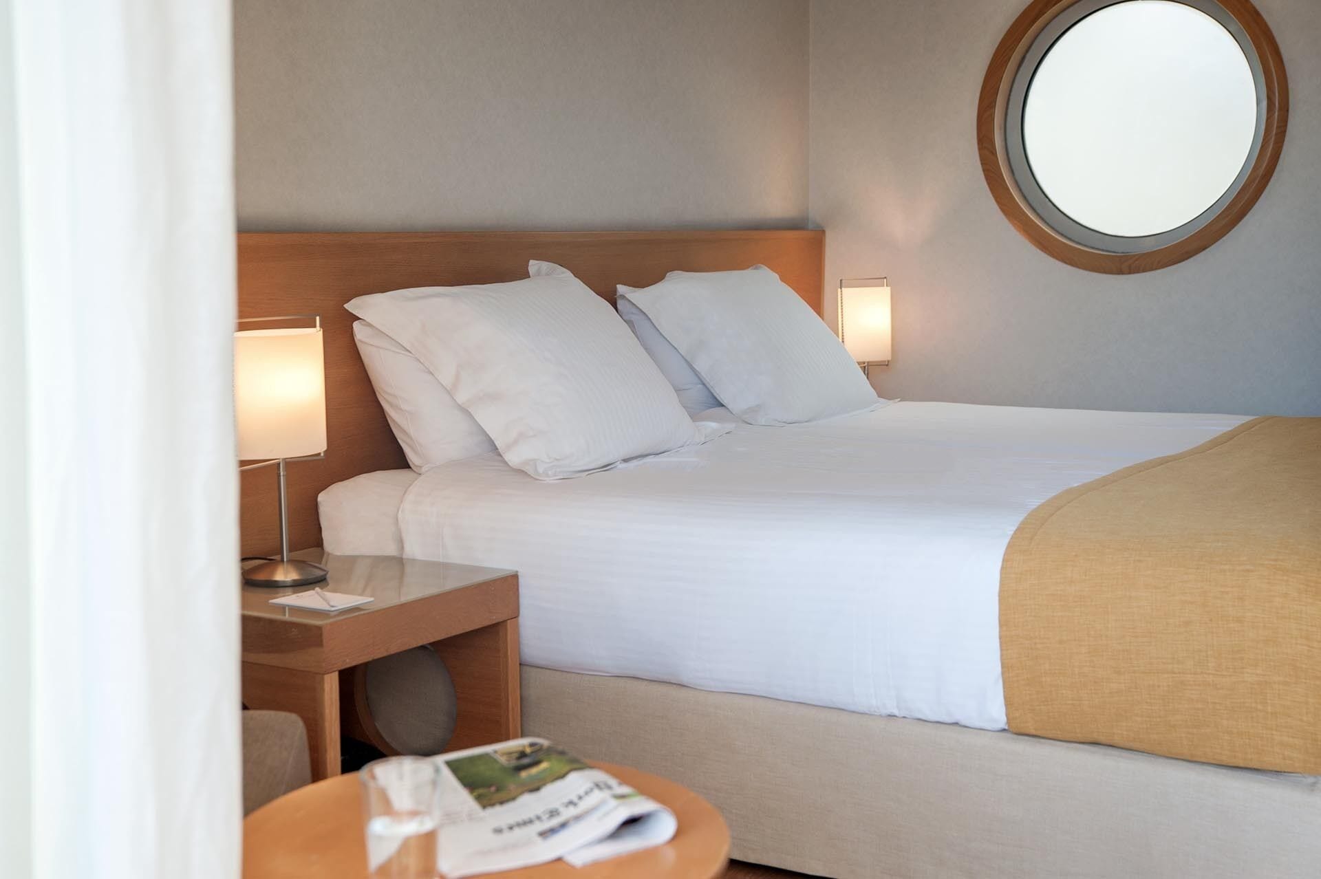 Habitación de hotel con cama, mesitas de noche, lámpara, espejo redondo y una pequeña mesa con un periódico.