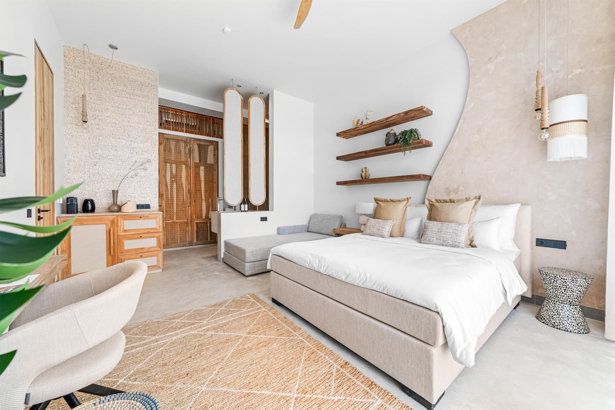 A modern bedroom with a large bed, a wooden dresser, a seating area, and a wall-mounted shelf with decorative items.