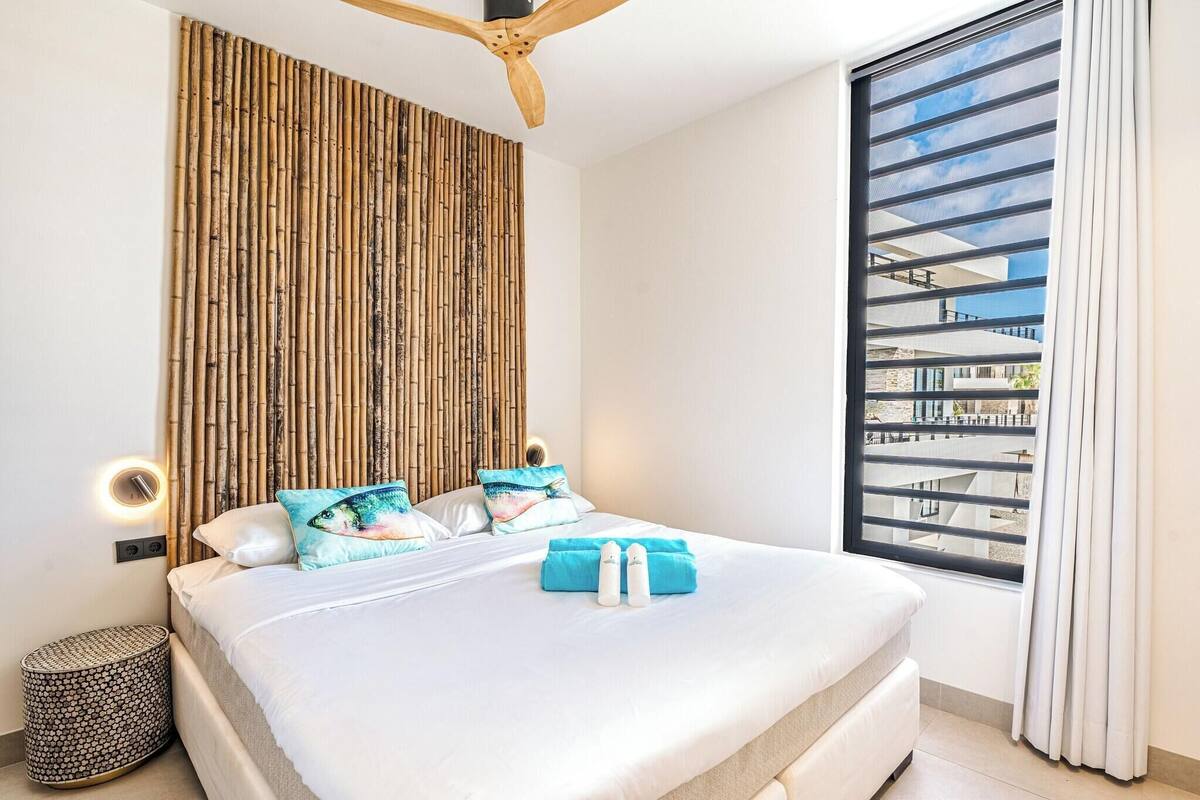 A modern hotel room with a large bed, a bamboo wall panel, a ceiling fan, a window with a view, and a bedside lamp.