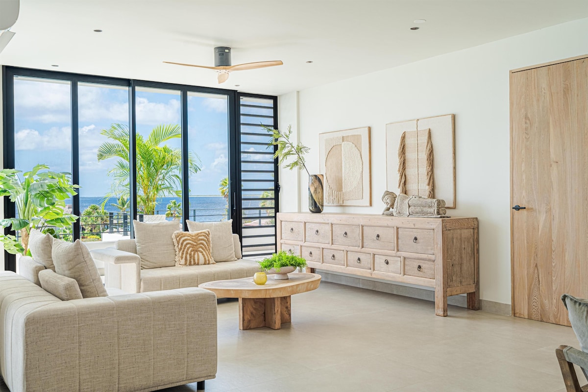 A modern living room with a large window, a ceiling fan, a wooden coffee table, and a beige sofa.