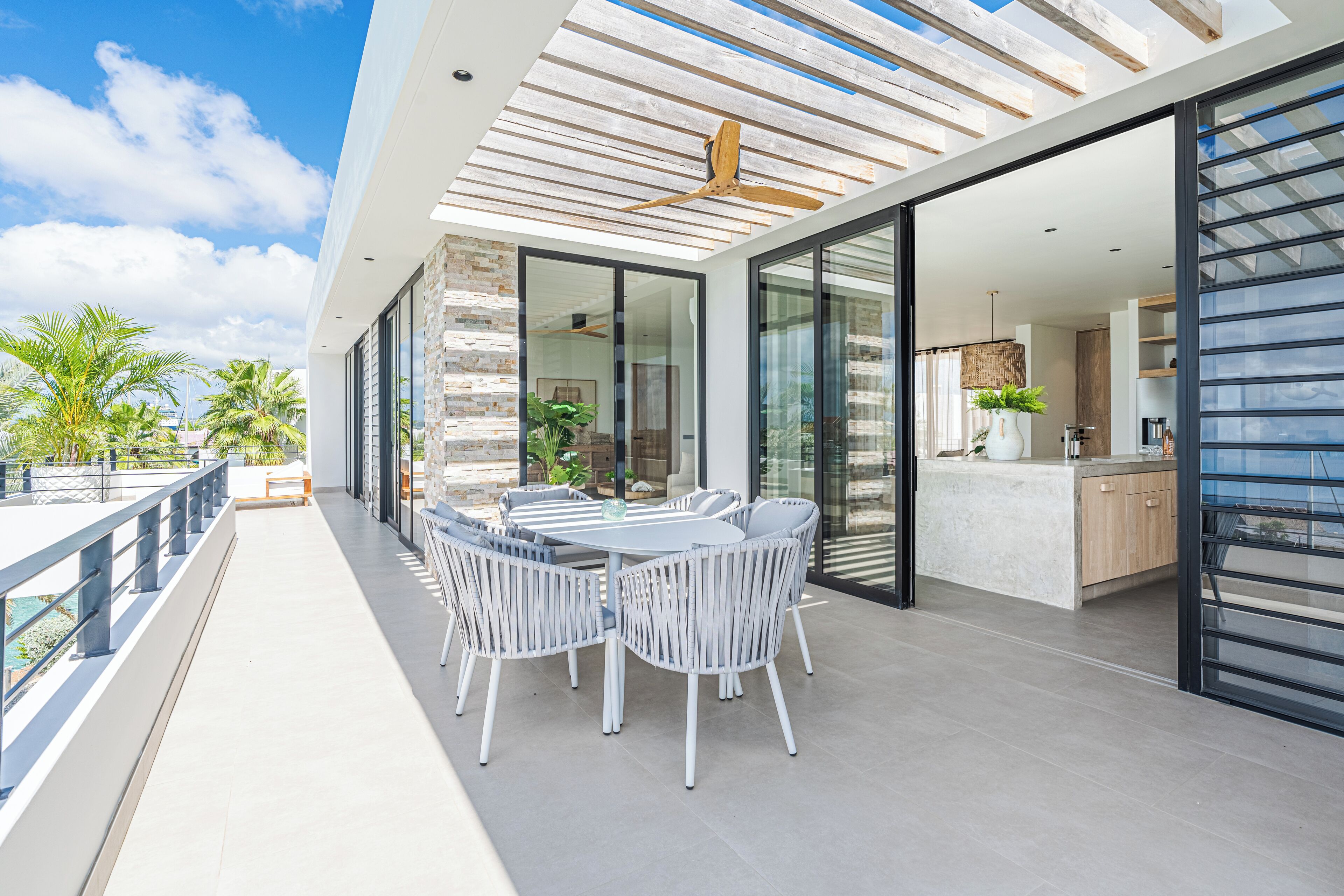 A modern outdoor dining area with a round table and white chairs, connected to a kitchen with a wooden countertop and a hanging plant.