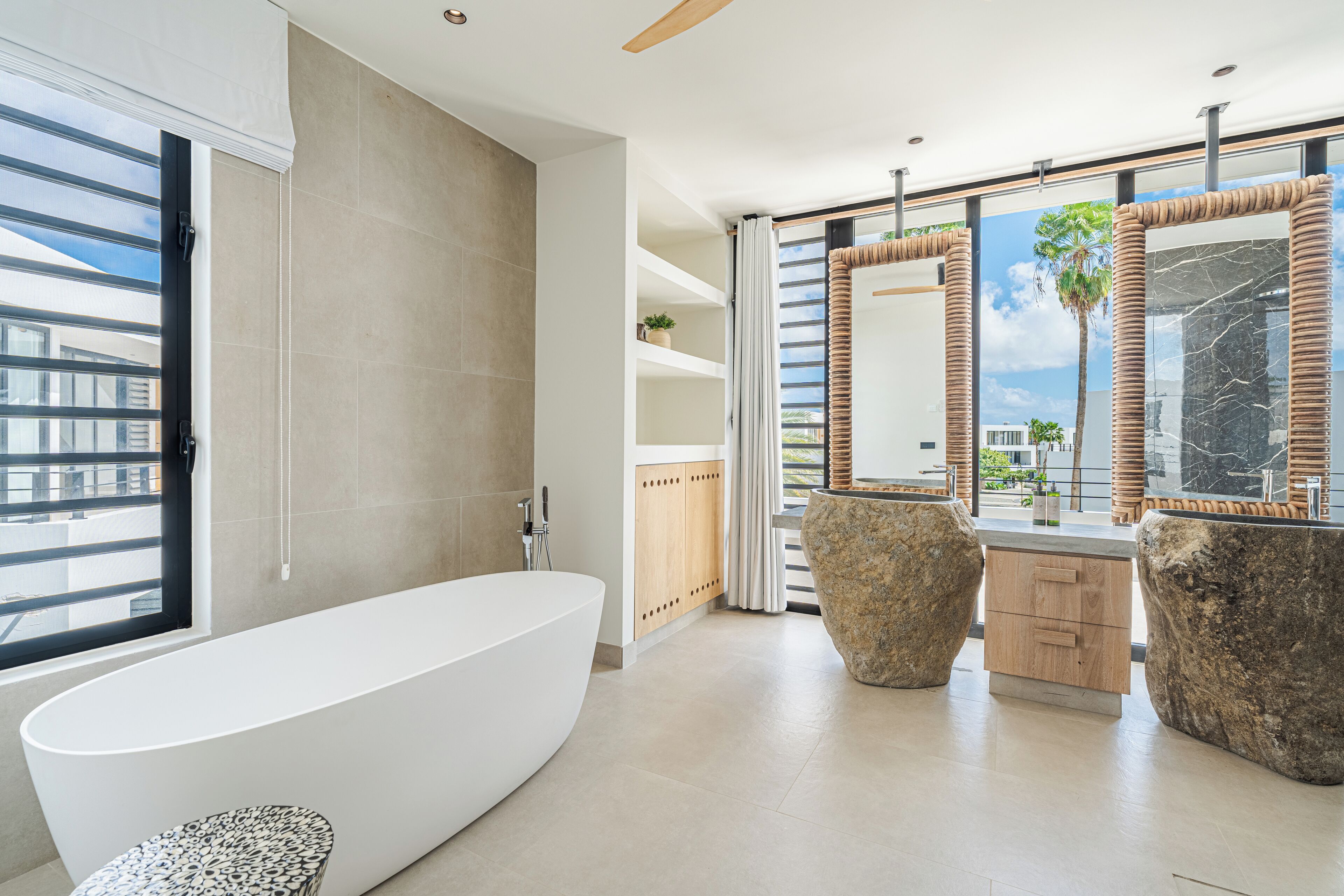 A modern bathroom with a freestanding bathtub, a large mirror, and a view of palm trees through the windows.