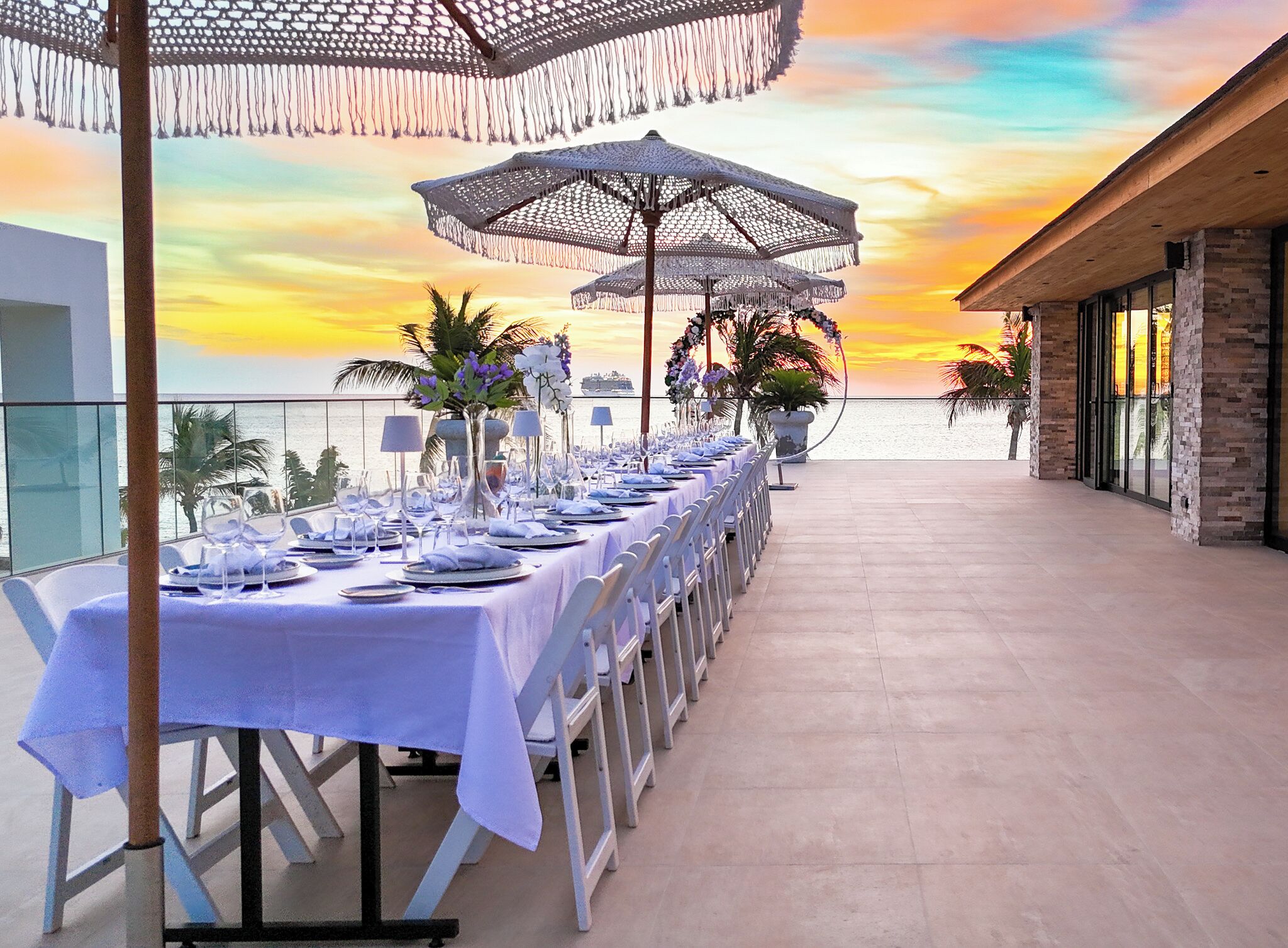 A long dining table set for a meal with white tablecloths and chairs, large umbrellas, and a view of the sunset over the ocean.