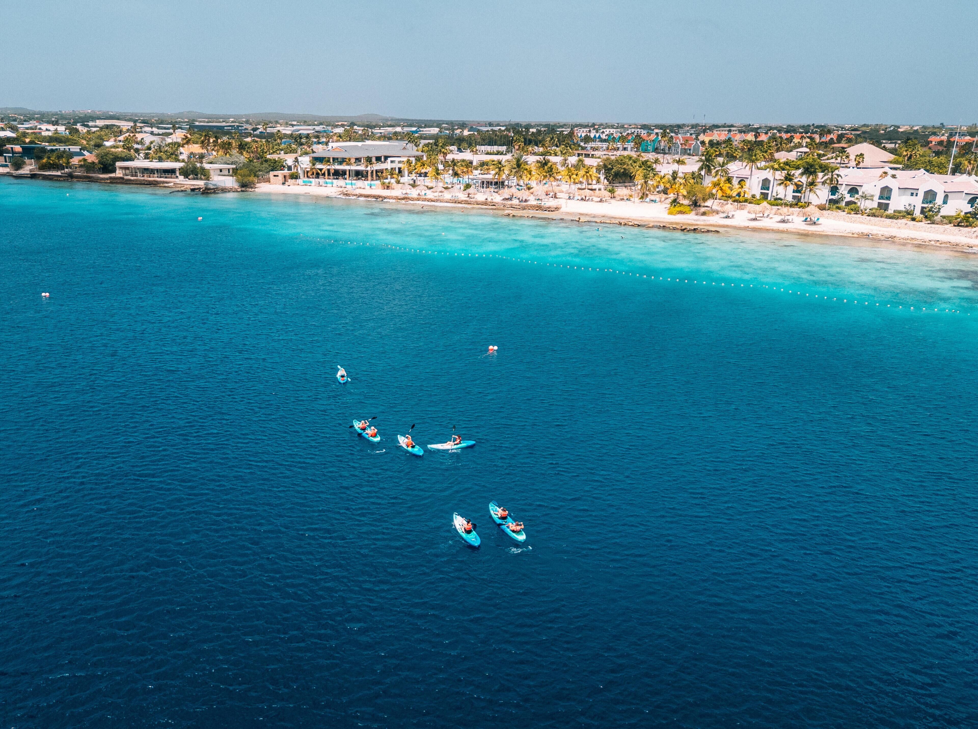 Vista aérea de una zona costera con aguas turquesas cristalinas, una playa de arena y una hilera de edificios a lo largo de la orilla.