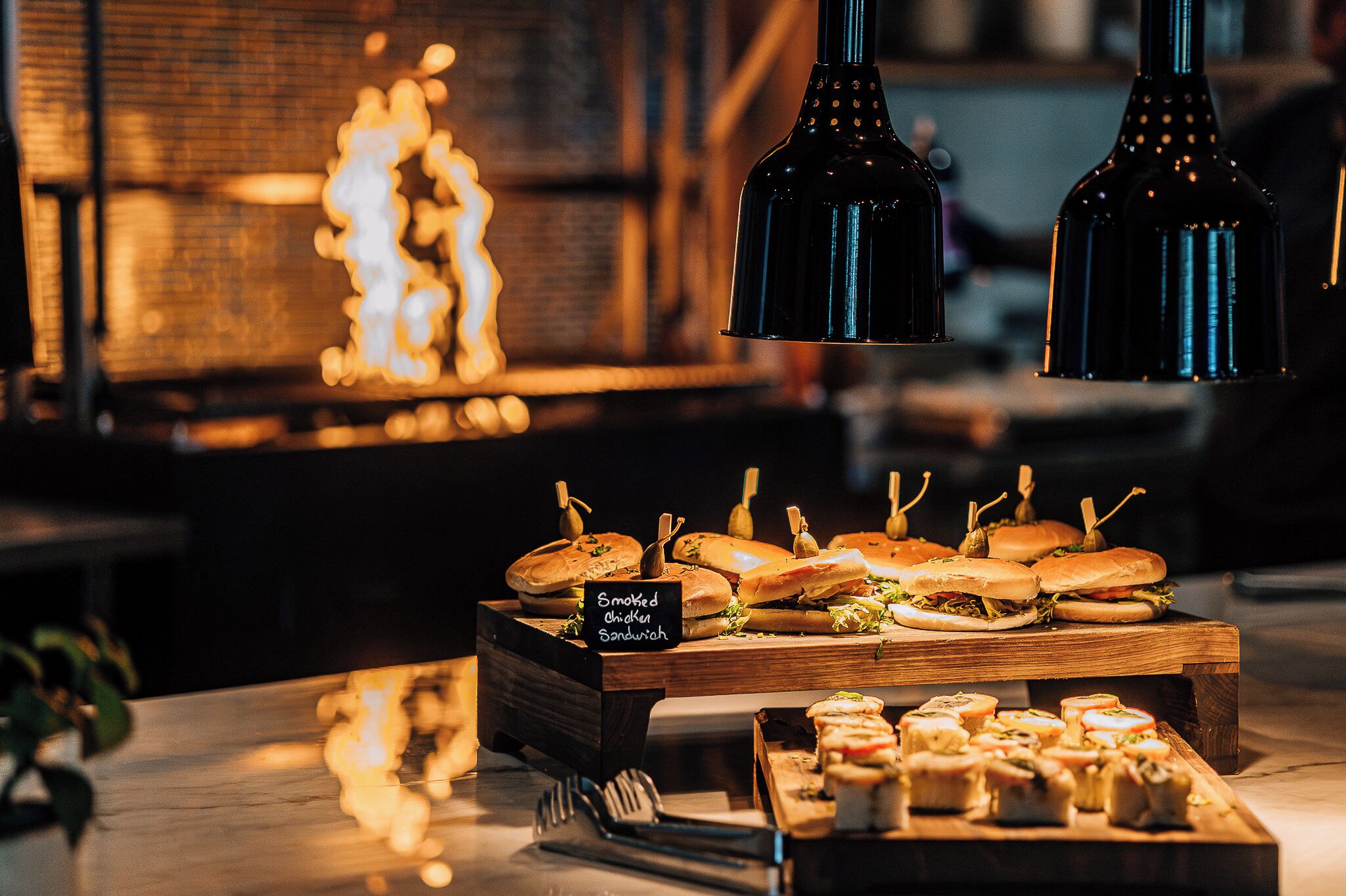 A buffet with sandwiches and appetizers, a sign indicating 'Smoked Chicken Sandwich', and hanging pendant lights.