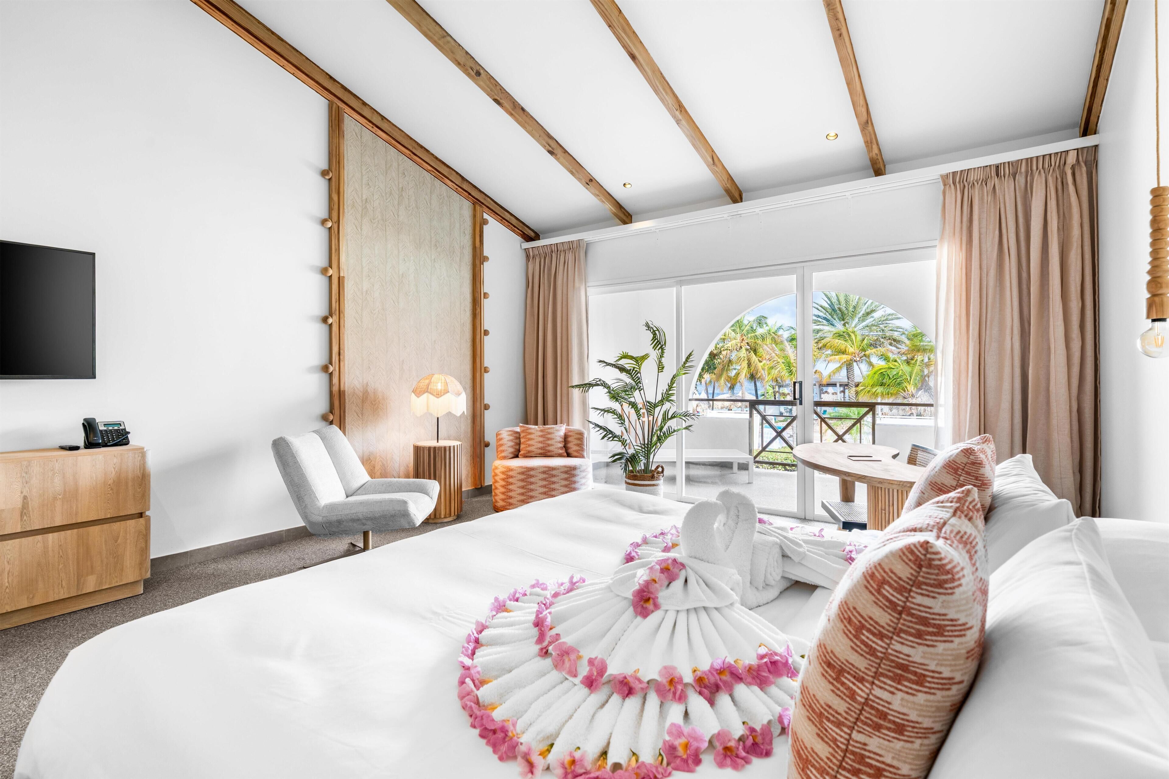 A hotel room with a large bed, a desk, a chair, a TV, and a view of palm trees.