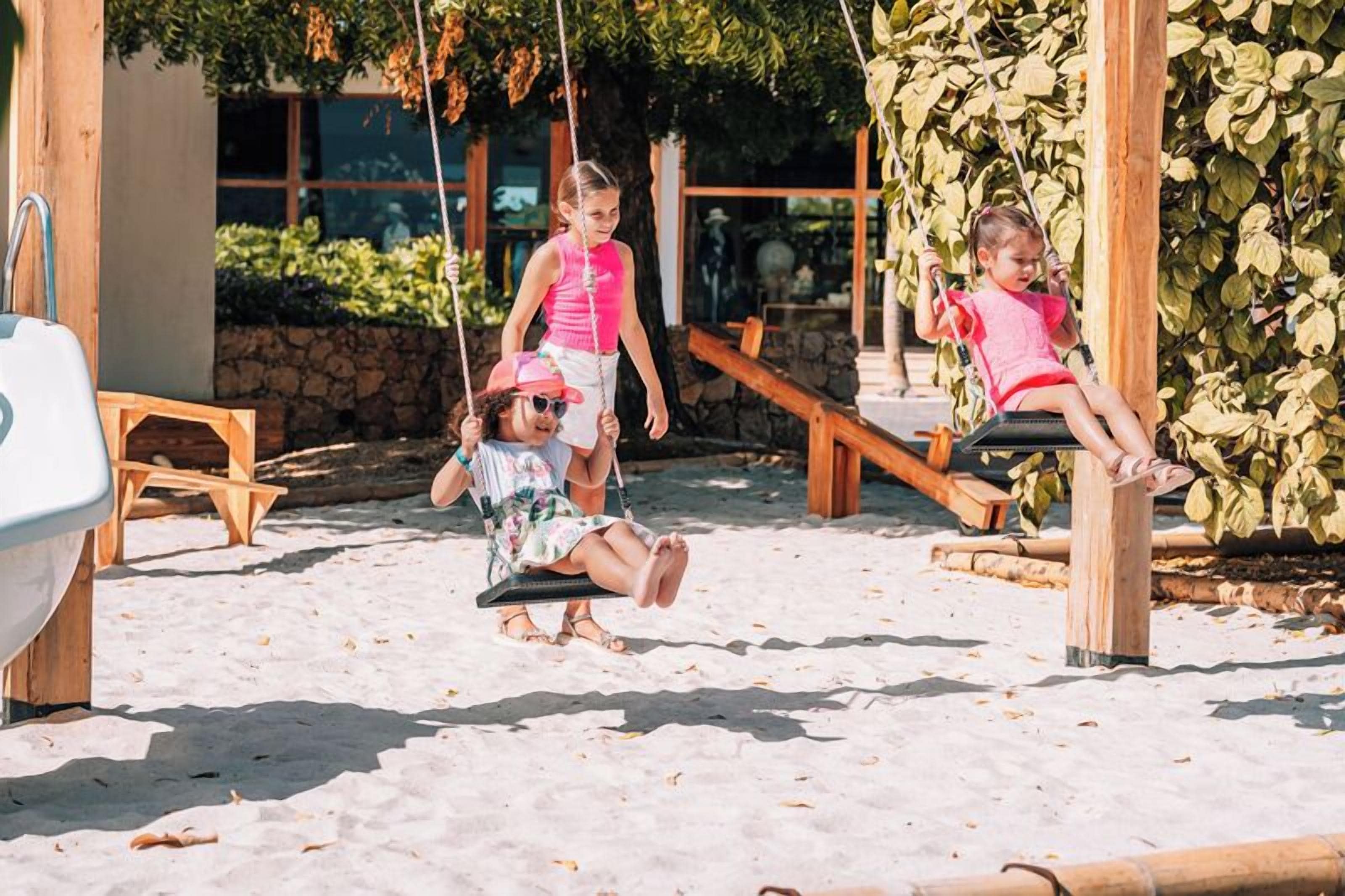 Children playing on swings in a sandy area with wooden structures and greenery in the background.