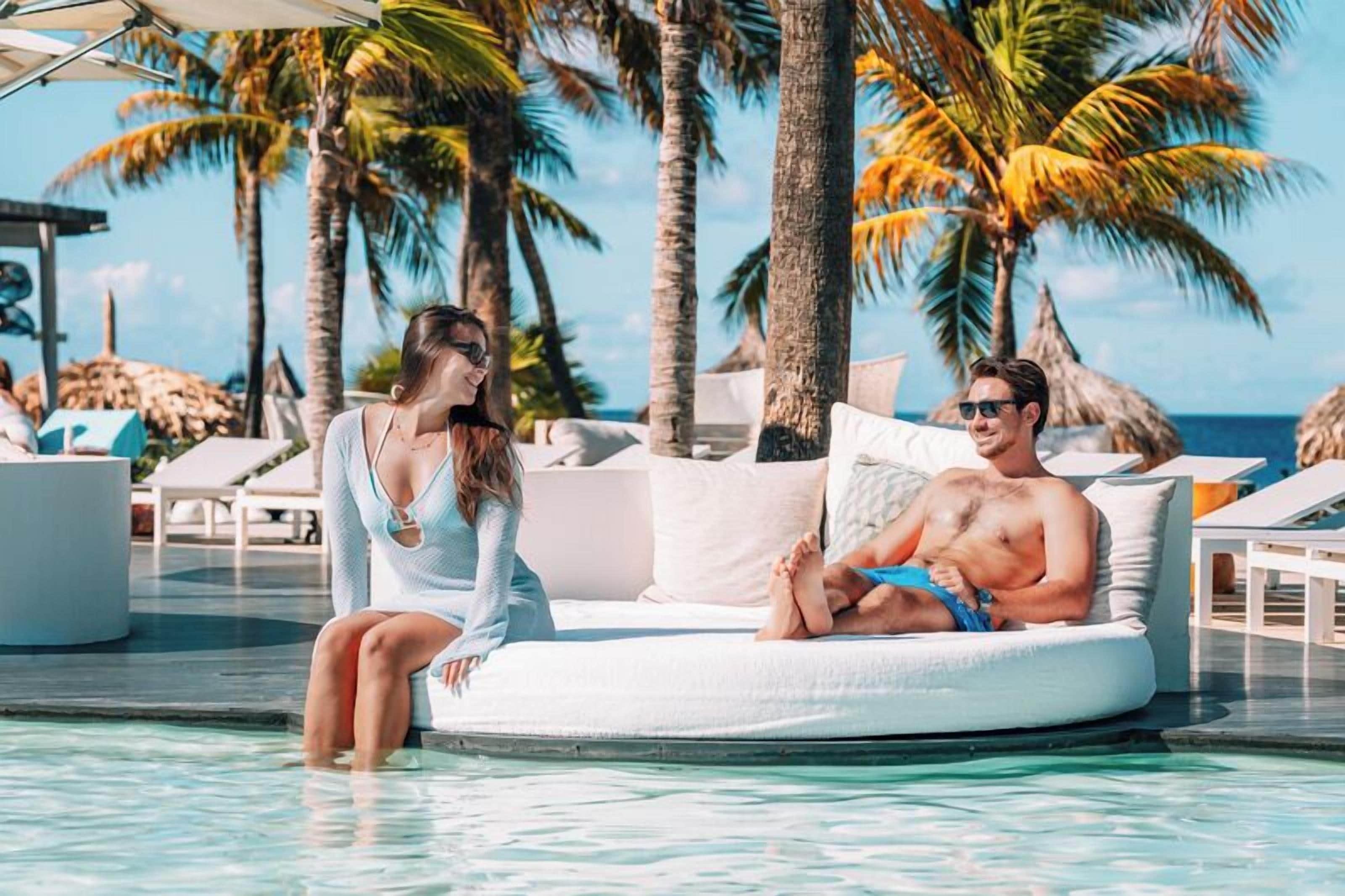A couple relaxing on an inflatable mattress in a pool area with lounge chairs and palm trees.
