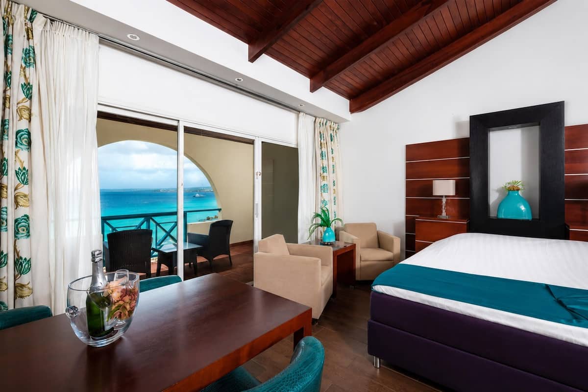 A hotel room with a bed, a wooden table, a chair, a sofa, a potted plant, and a view of the sea.