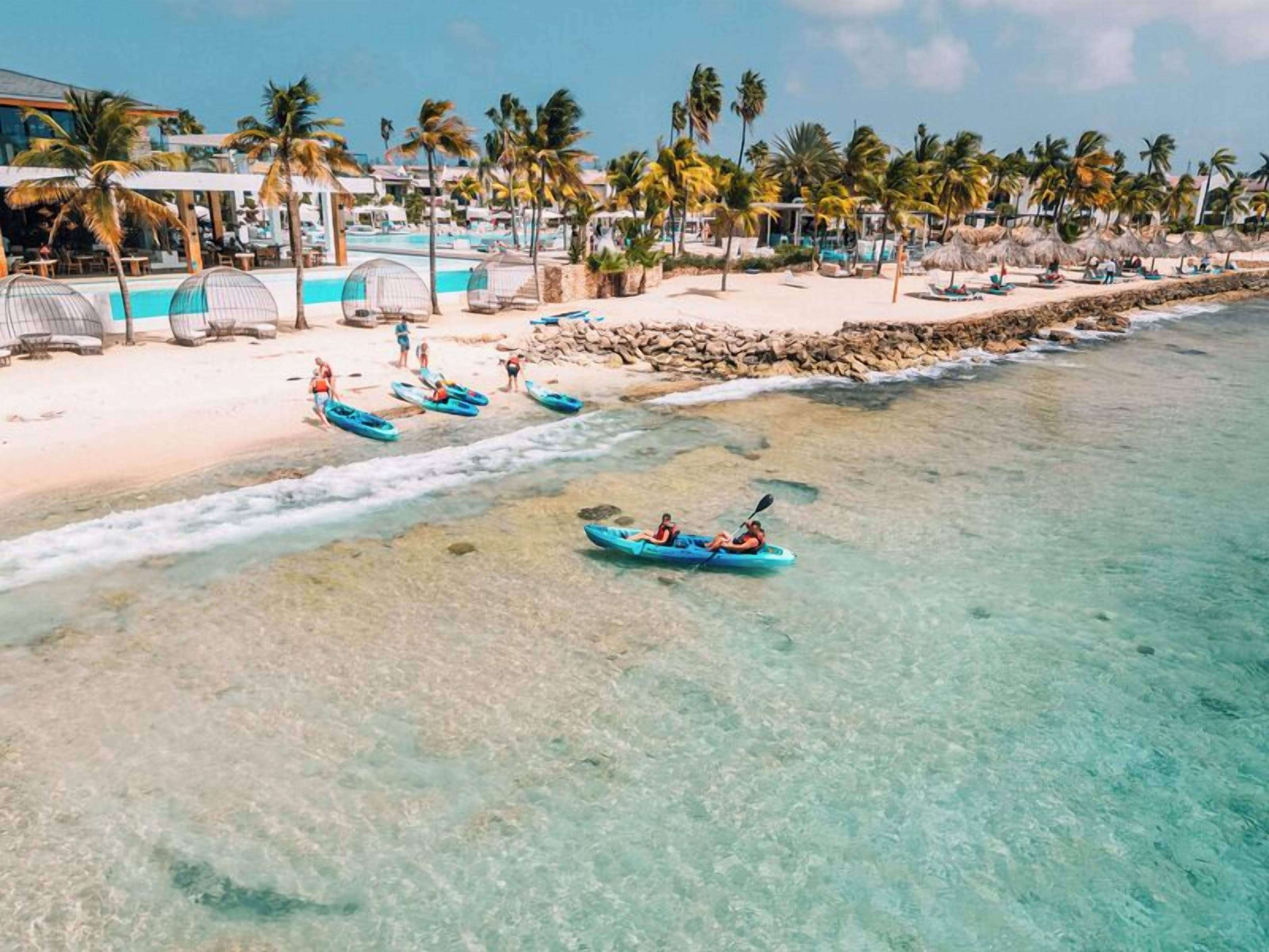 A beach resort with clear turquoise waters, kayaks, and palm trees.