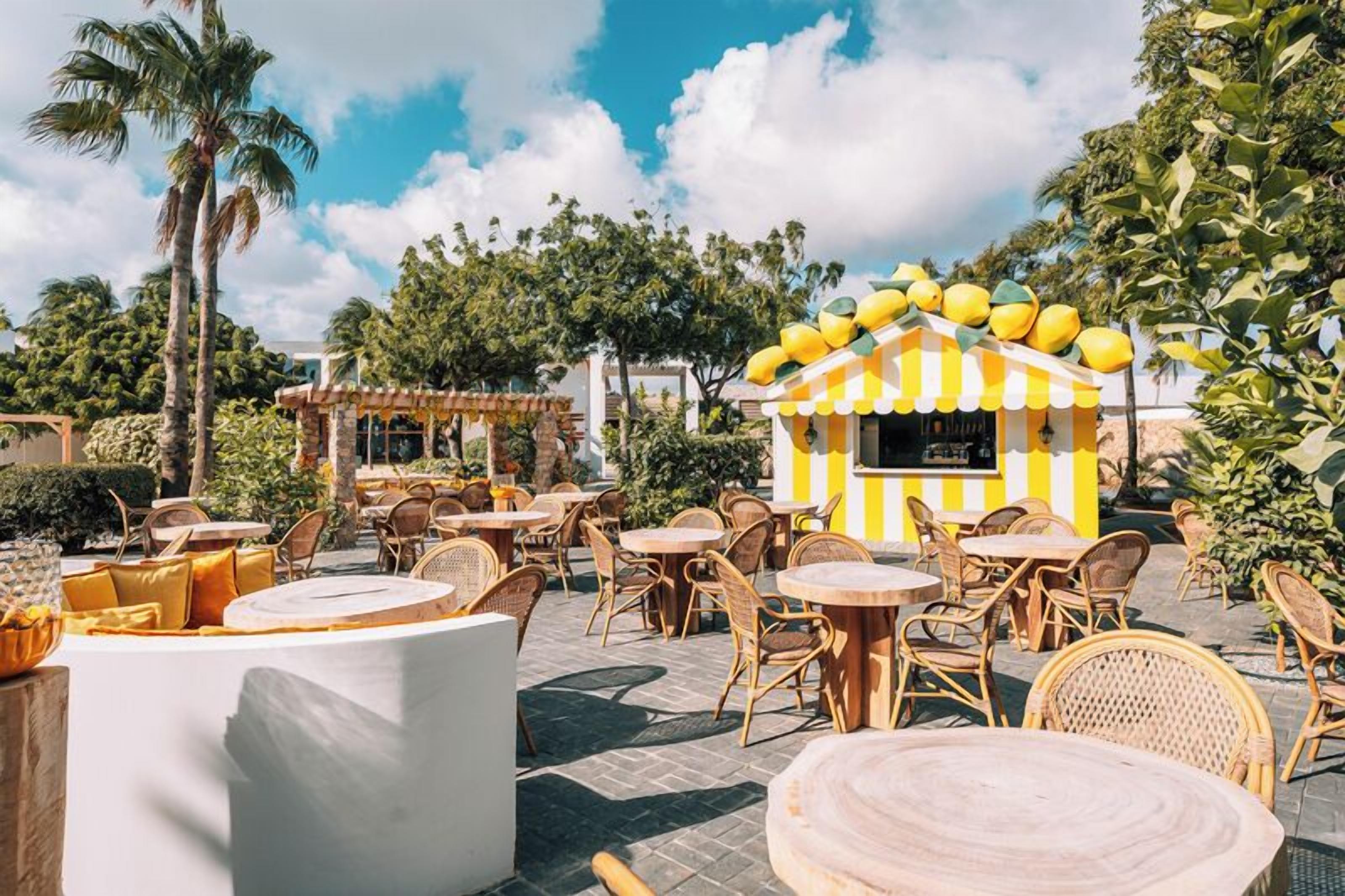 An outdoor seating area with round tables and wicker chairs, a small yellow and white striped kiosk, and palm trees in the background.