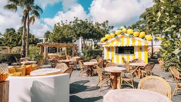 An outdoor seating area with round tables and wicker chairs, a small yellow and white striped kiosk, and palm trees in the background.