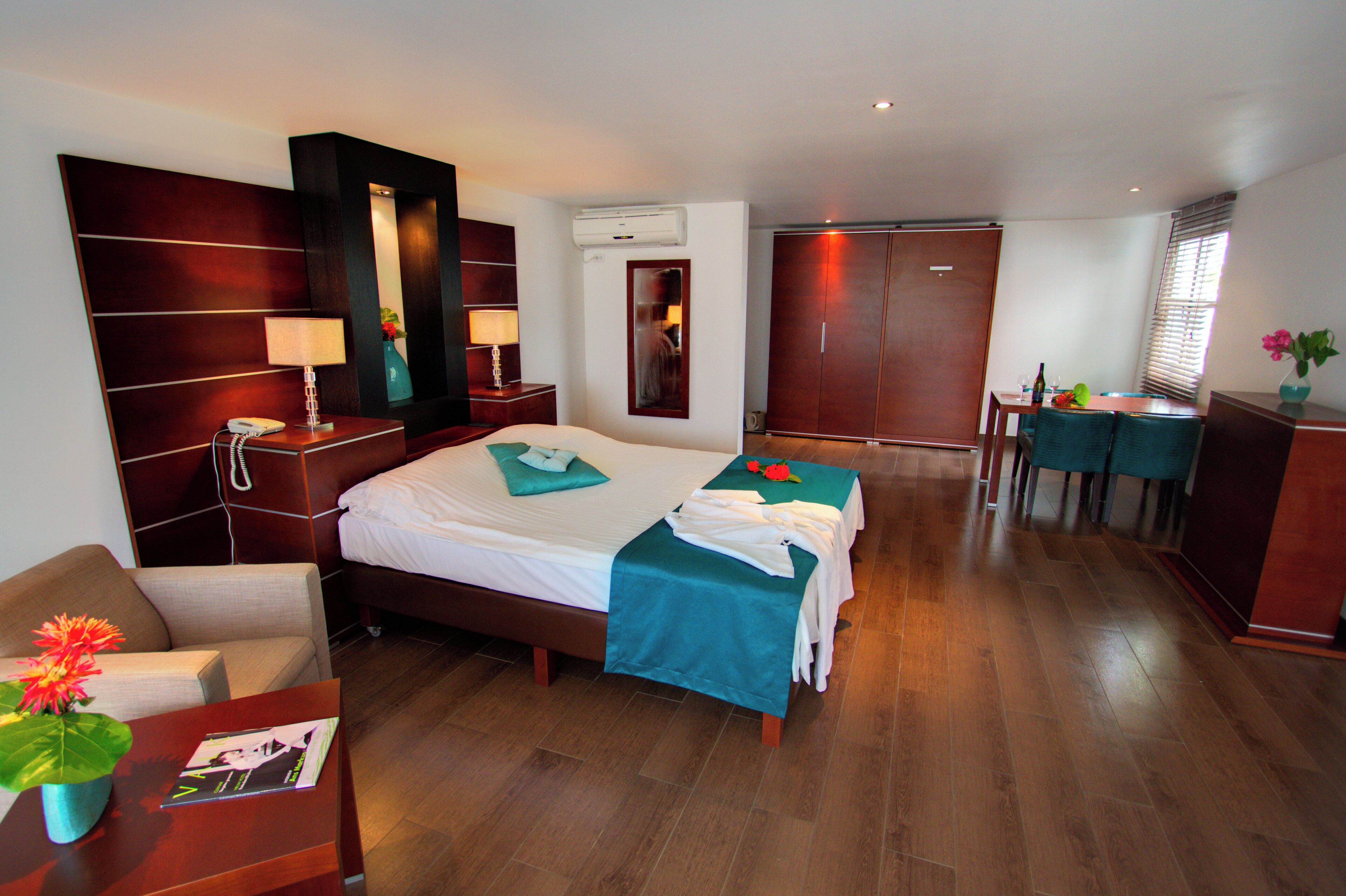 A hotel room with a bed, bedside table, lamp, mirror, wardrobe, and a small desk with a chair.