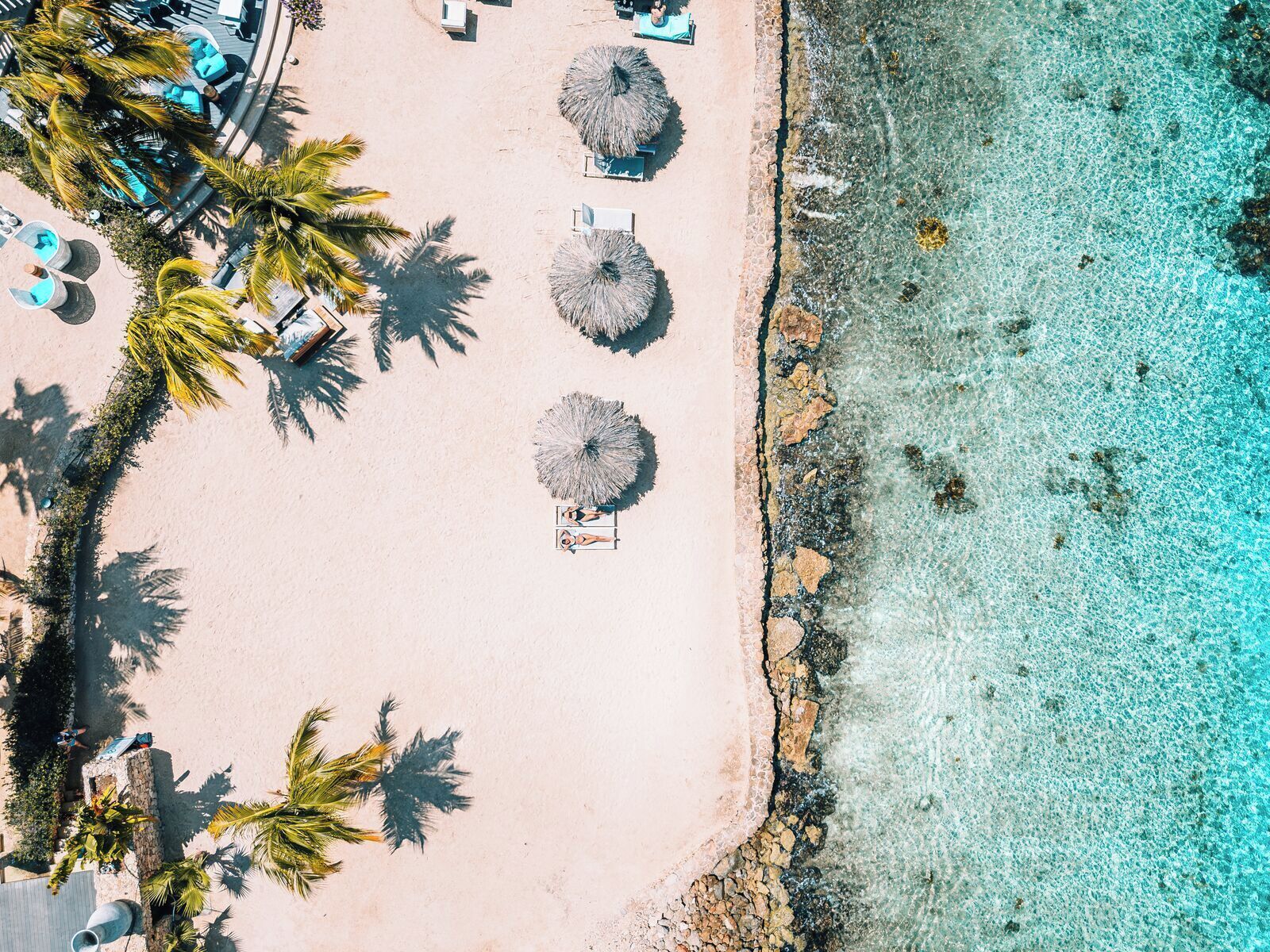 Eine Luftaufnahme eines Strandes mit klarem türkisblauem Wasser, Palmen und Liegestühlen.
