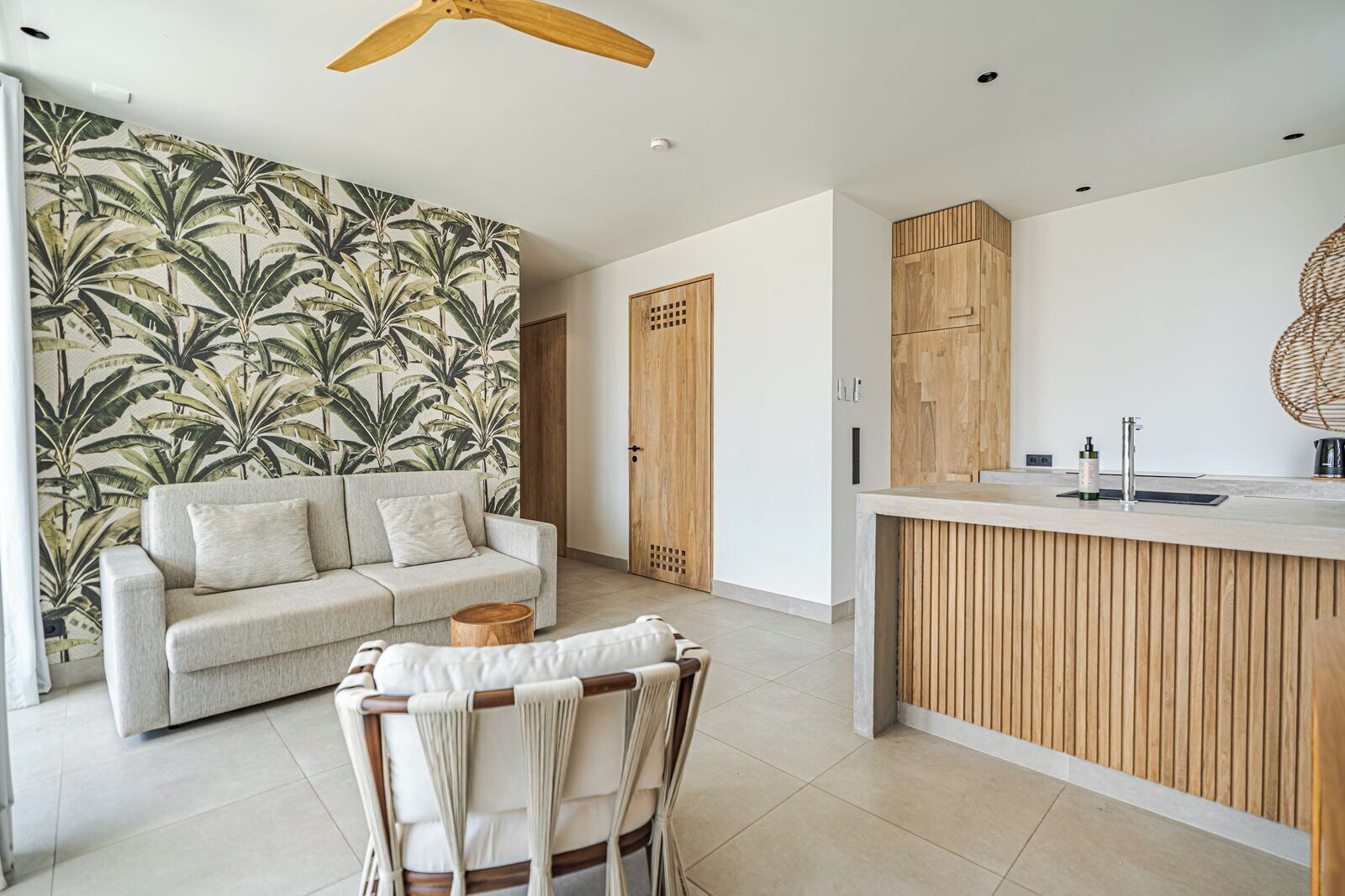 Una sala de estar moderna con un sofá, una silla de madera, una mesita redonda y una zona de cocina con encimera y armarios de madera.