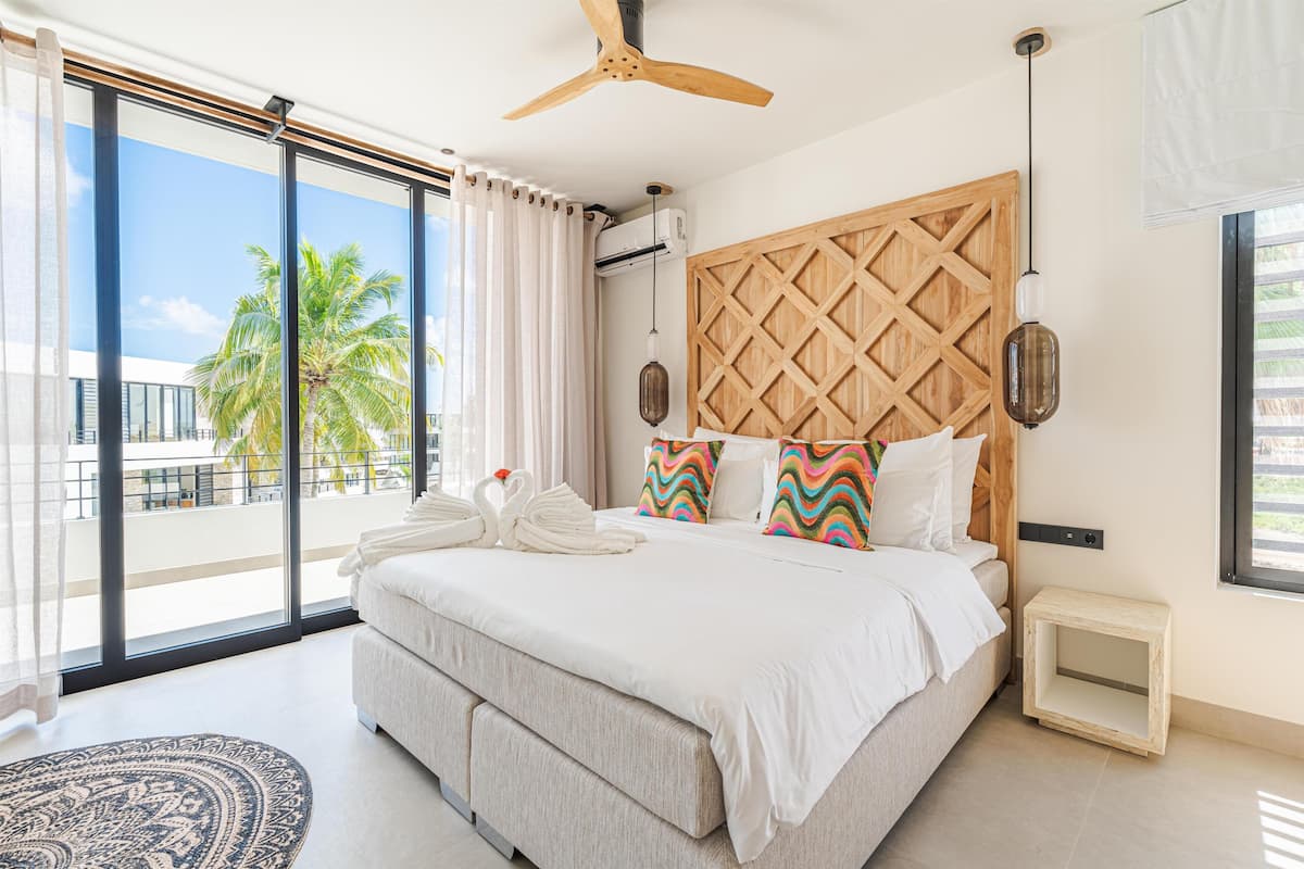 A modern bedroom with a large bed, a ceiling fan, a wooden headboard, and a view of palm trees through large windows.