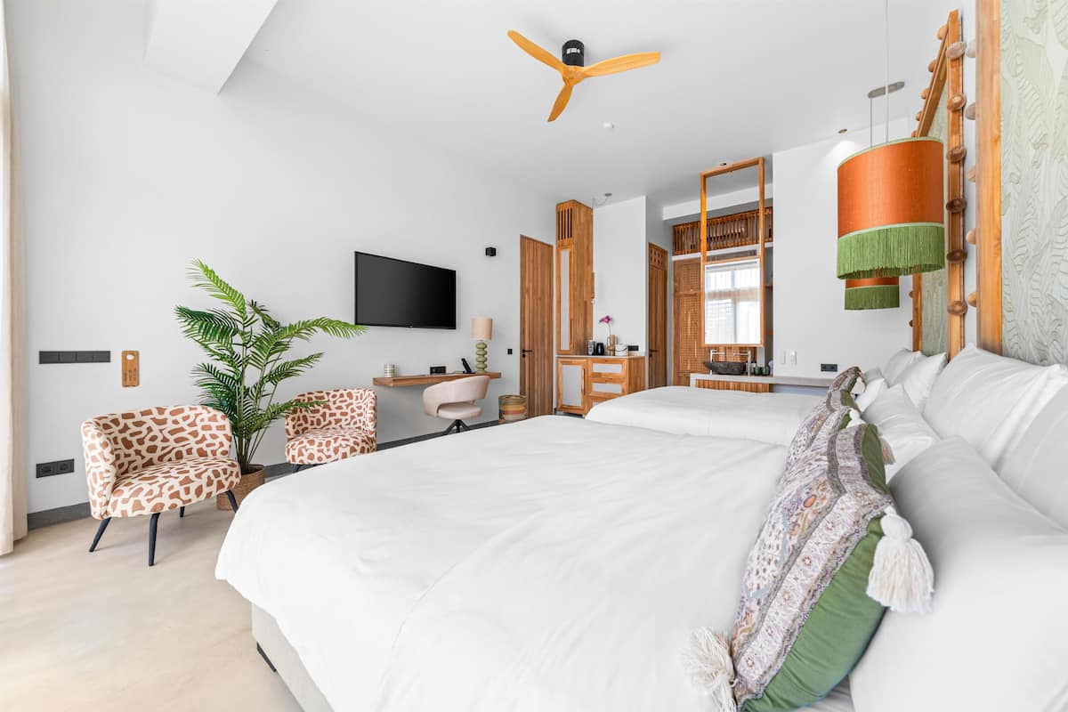 A modern hotel room with a large bed, a ceiling fan, a TV, a potted plant, and a seating area with a chair and a small table.