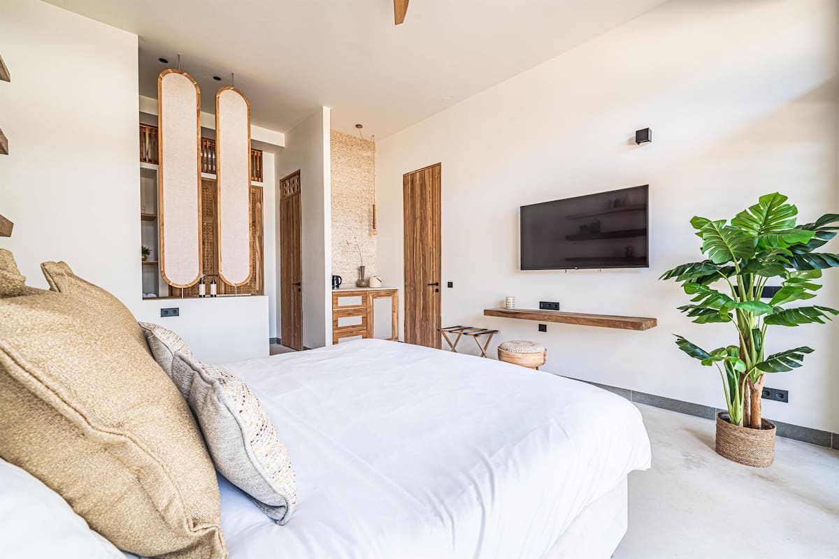 A modern bedroom with a large bed, a TV mounted on the wall, a plant, and a wooden cabinet.