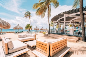 Un salon de plage doté de mobilier en bois, de parasols en chaume et d’un espace piscine.