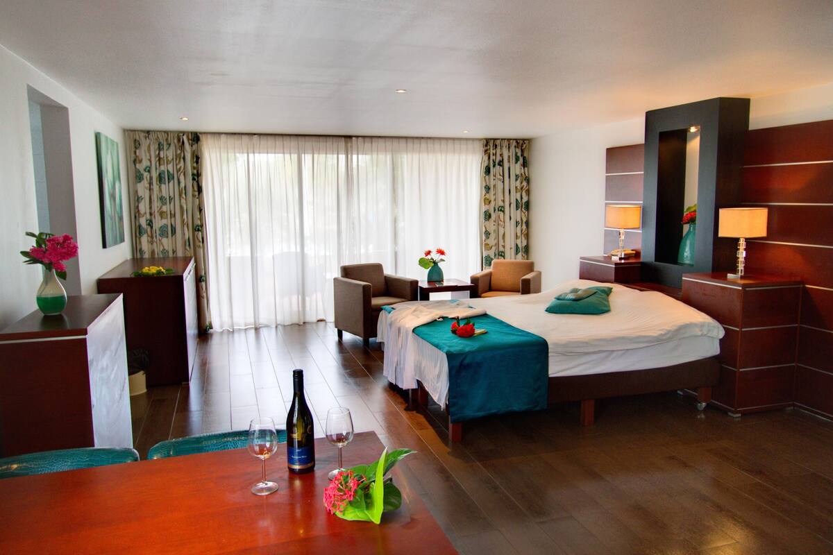 A hotel room with a bed, two chairs, a desk, a mirror, and a table with a bottle and glass.