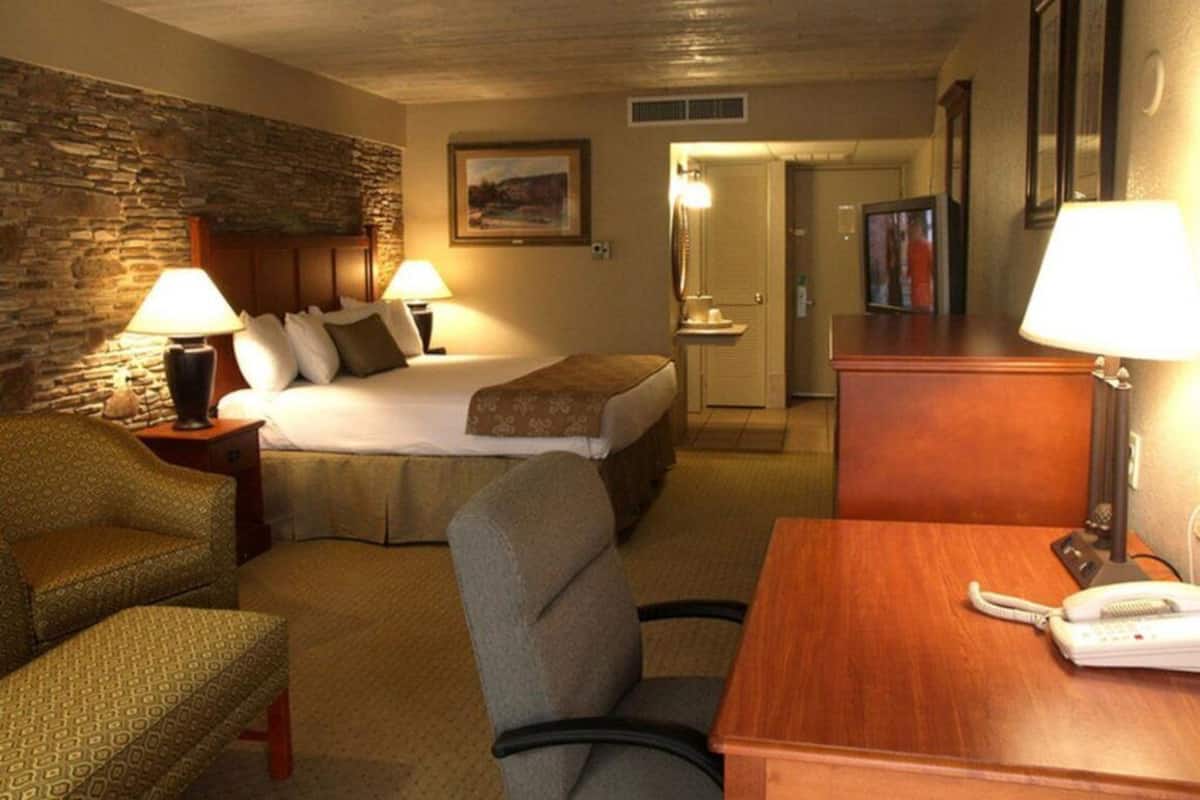 A hotel room with a bed, two bedside lamps, a desk with a telephone, a chair, and a stone wall.