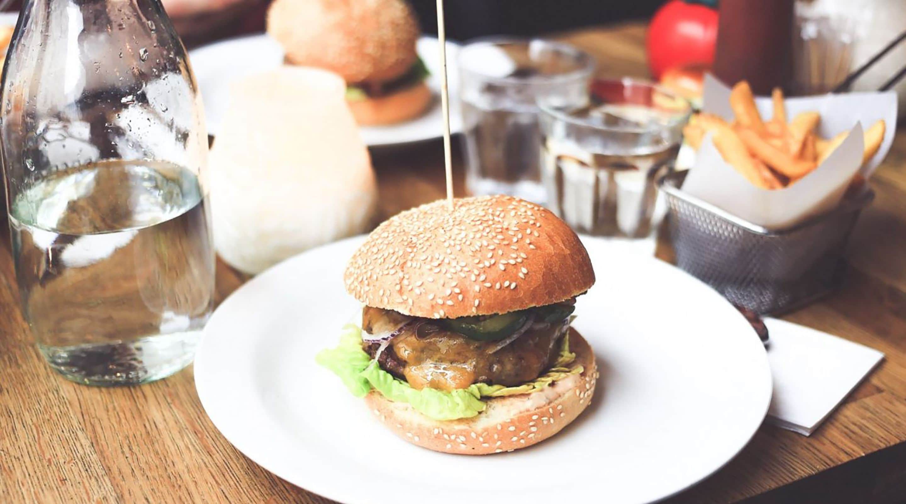 A burger on a white plate with a bottle of water and fries in the background.