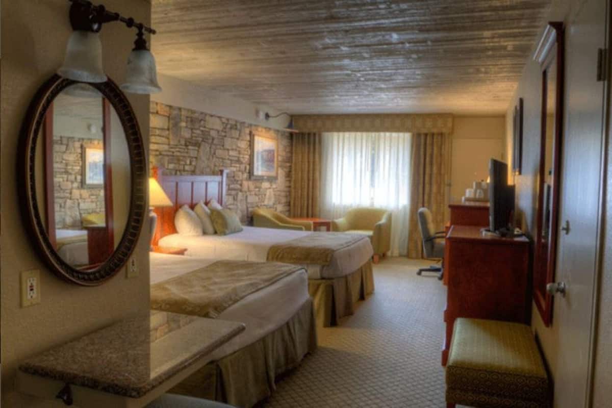 A hotel room with two beds, a stone wall, a large mirror, a television, and a desk.