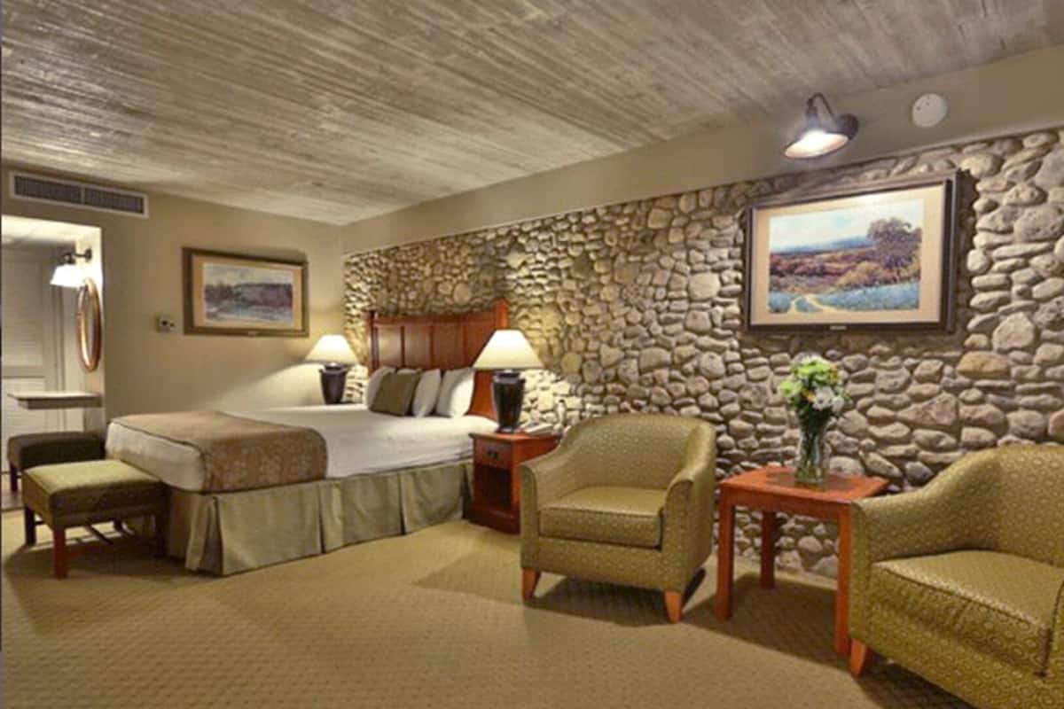 A hotel room with a stone wall, a bed, two chairs, a small table with a vase, and two lamps.