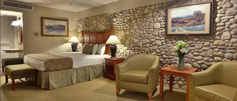 A hotel room with a stone wall, a bed, two chairs, a small table with a vase, and two lamps.