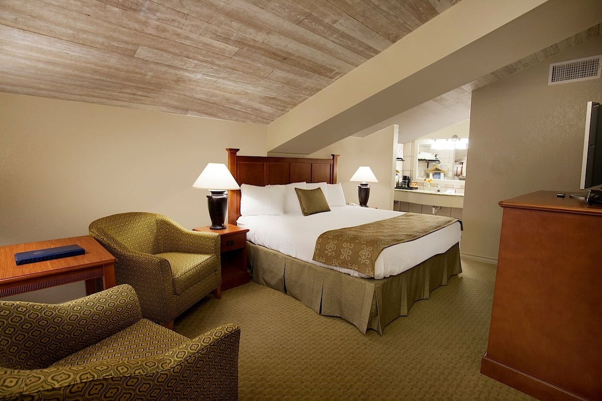 A hotel room with a bed, two bedside lamps, a wooden nightstand, a patterned armchair, and a desk with a lamp.