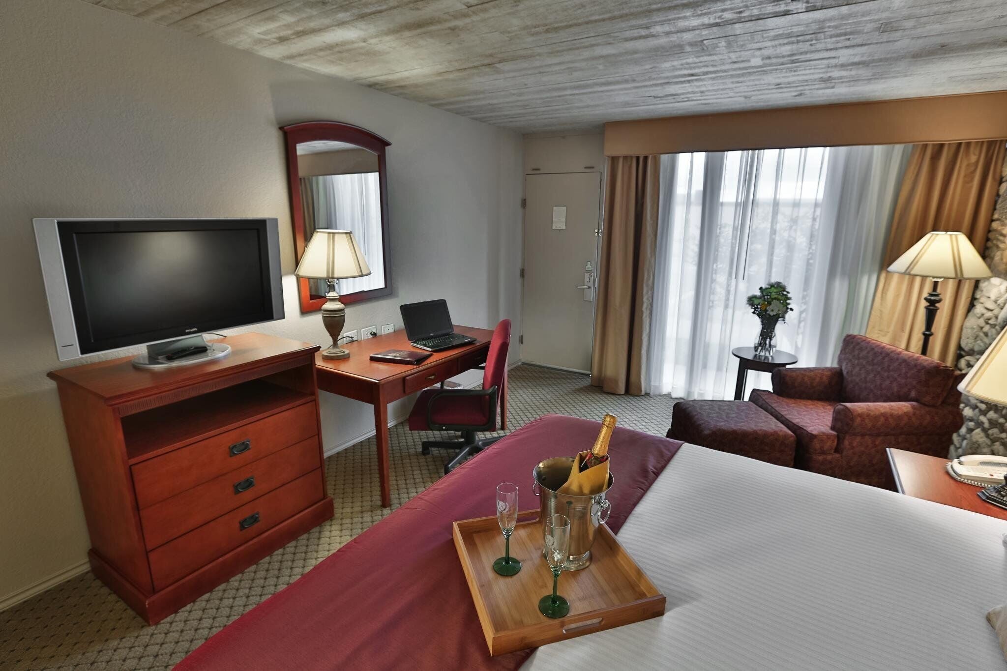Hotellihuone, jossa on sänky, työpöytä tietokoneella, televisio, minibaari ja ikkuna, jossa on verhot.