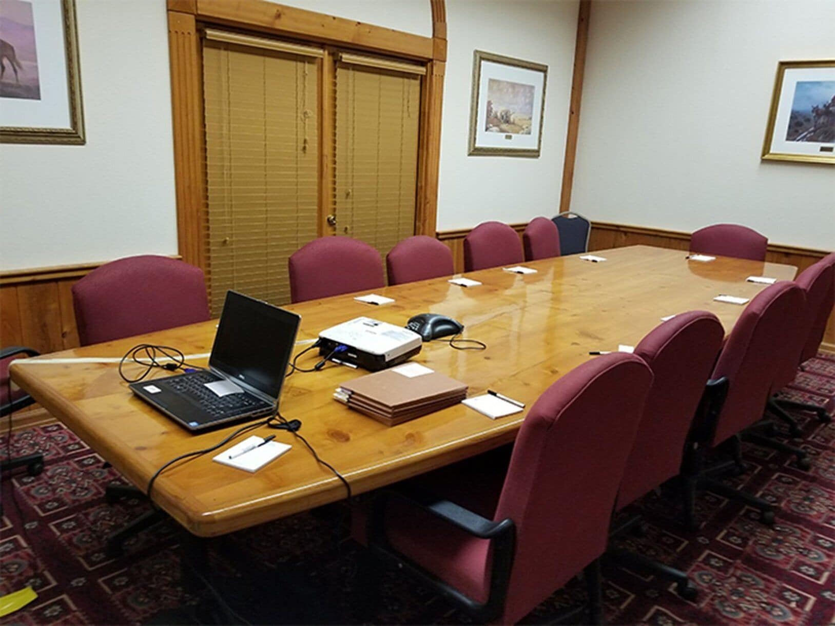 A conference room with a wooden table, red chairs, a laptop, a projector, and a whiteboard.