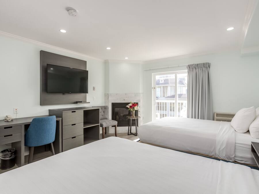 Quarto de hotel com duas camas, uma escrivaninha com cadeira, televisão de tela plana na parede, lareira e janela com cortinas.