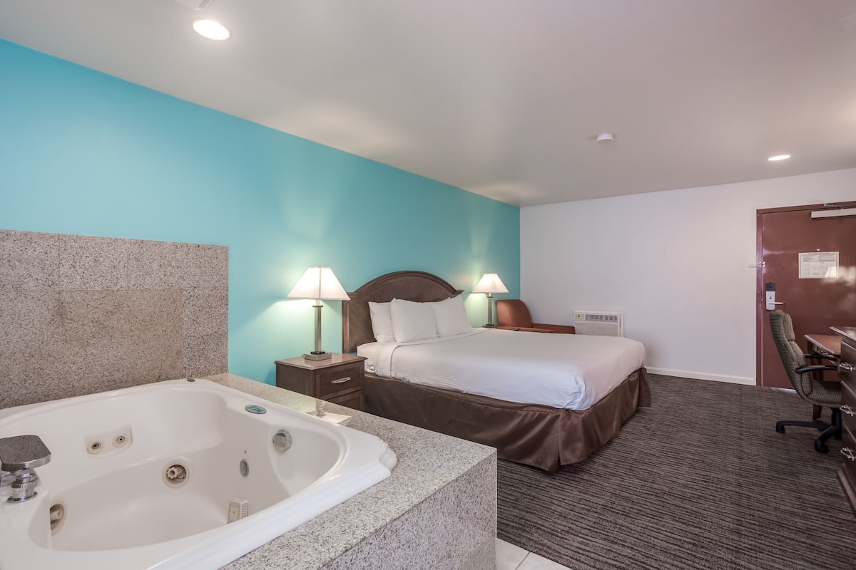 A hotel room with a large bathtub, a bed with a brown headboard, two bedside lamps, and a desk with a chair.