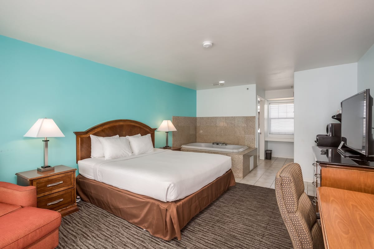 A hotel room with a bed, two bedside lamps, a TV, a desk, and a bathroom with a bathtub.