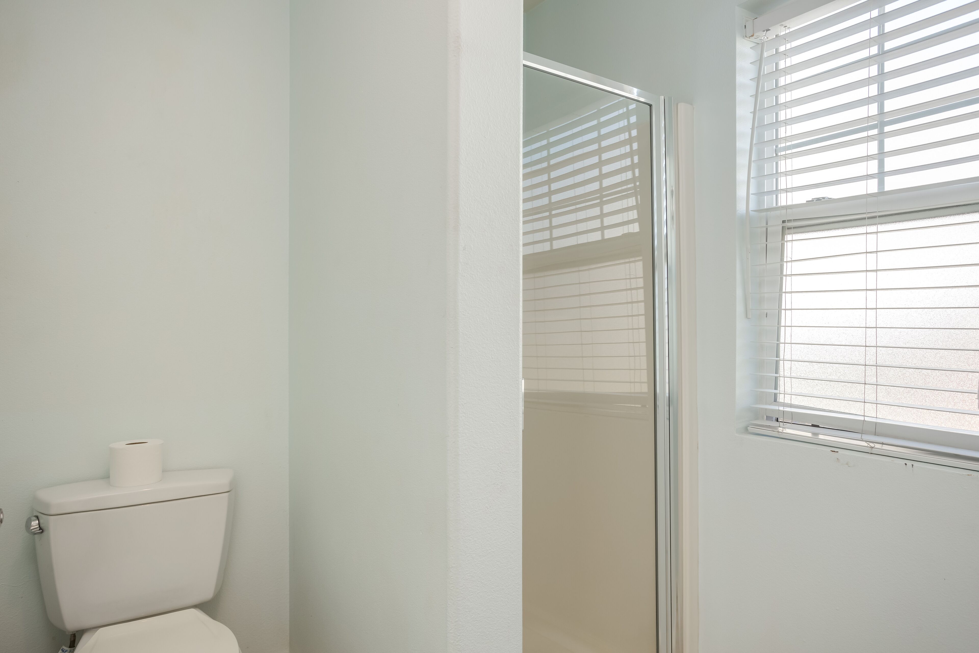 A bathroom with a toilet, a glass shower enclosure, and a window with blinds.
