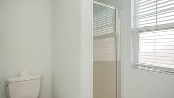 A bathroom with a toilet, a glass shower enclosure, and a window with blinds.
