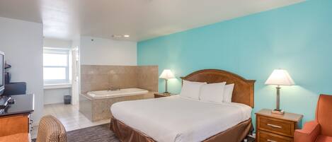 A hotel room with a large bed, two bedside lamps, a wooden nightstand, a desk with a chair, a bathtub, and a window with natural light.