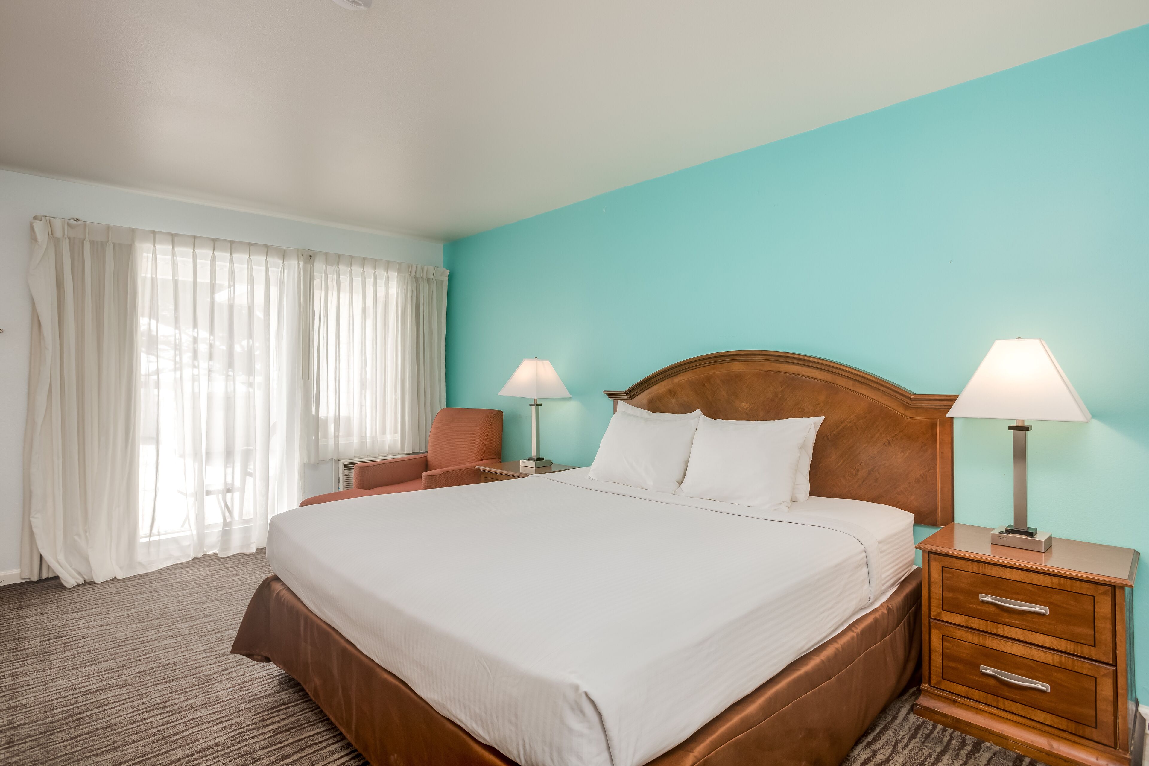 Habitación de hotel con una cama grande, dos mesitas de noche con lámparas, una silla y una ventana con cortinas.