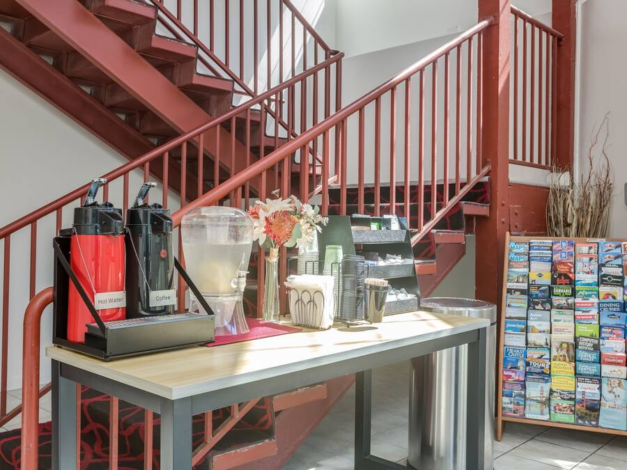 Uma escadaria com guarda-corpos vermelhos, uma mesa com dispensadores de café e água quente, e um painel de informações turísticas.