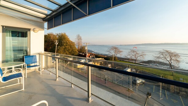 A balcony with a glass railing overlooking a body of water, a table with chairs, and a view of the shoreline.