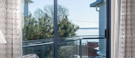 A living room with a patterned sofa, a wooden coffee table, and a large sliding glass door leading to a balcony with a view of the sea and buildings.