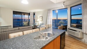 A modern kitchen with a granite countertop, stainless steel sink, and a view of the sunset through large windows.