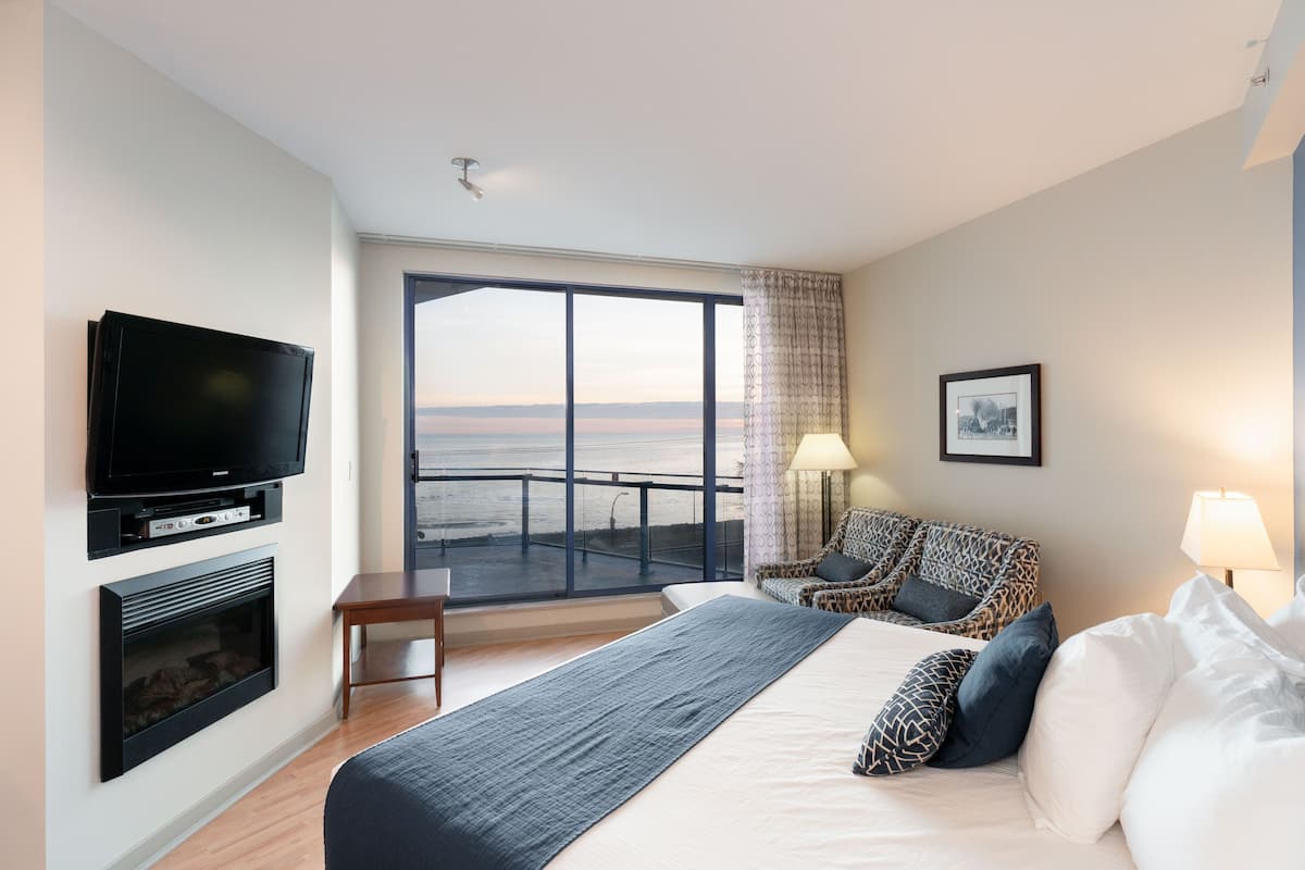 A hotel room with a large bed, a TV, a fireplace, a balcony with a view, and a patterned armchair.