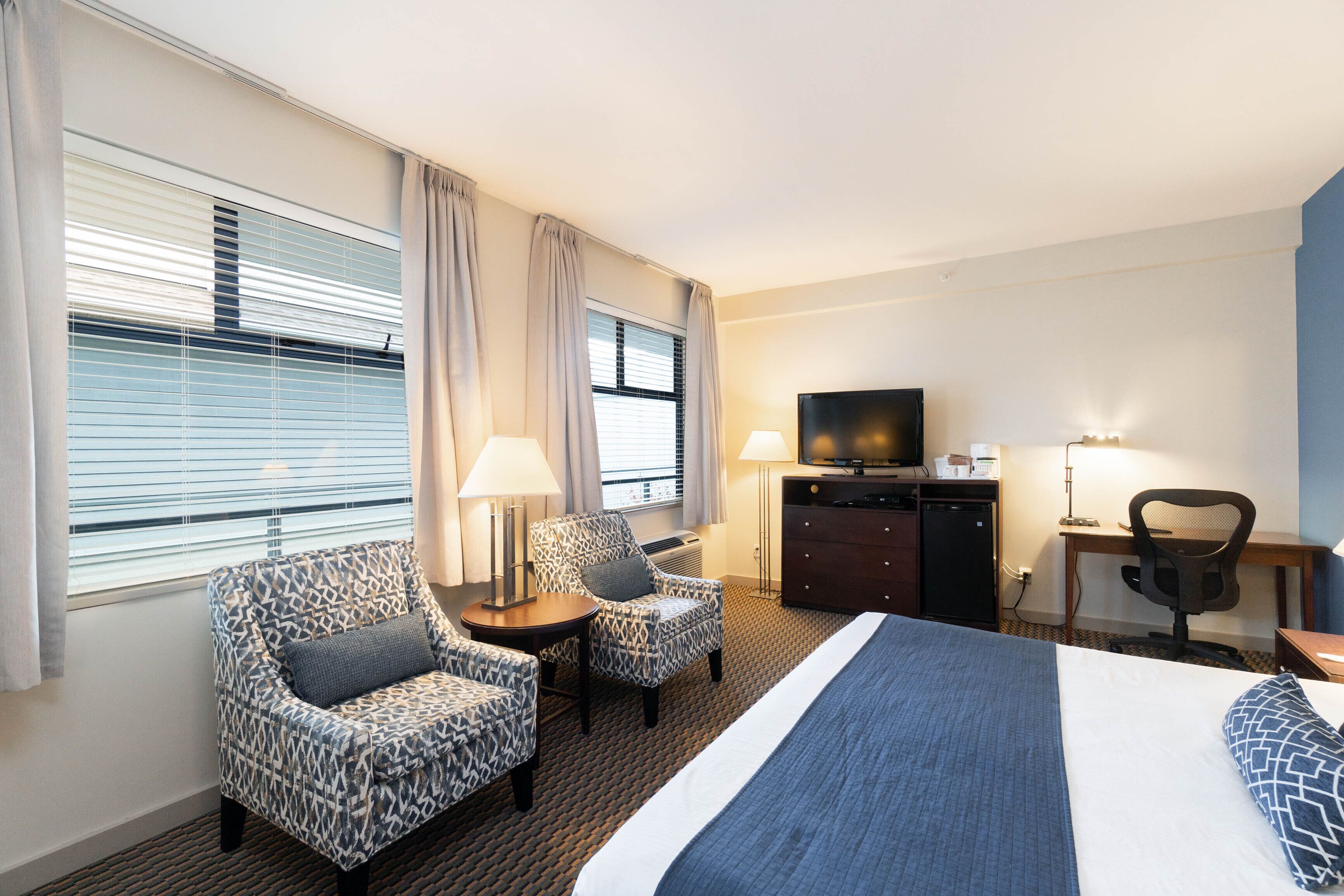 A hotel room with a large bed, two patterned armchairs, a desk with a television, and a dresser.