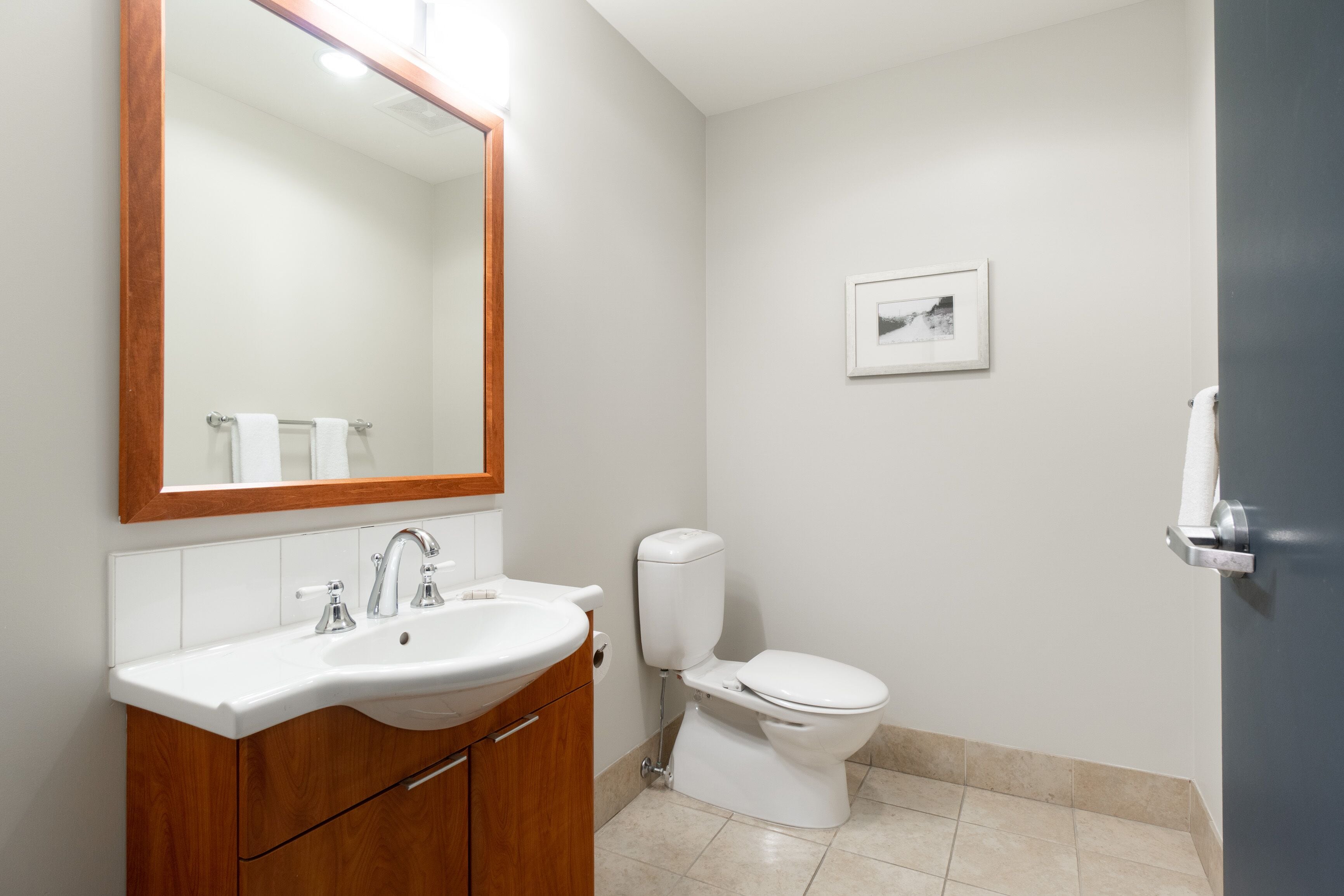 A bathroom with a wooden vanity, a white sink, a mirror, a toilet, and a framed picture on the wall.