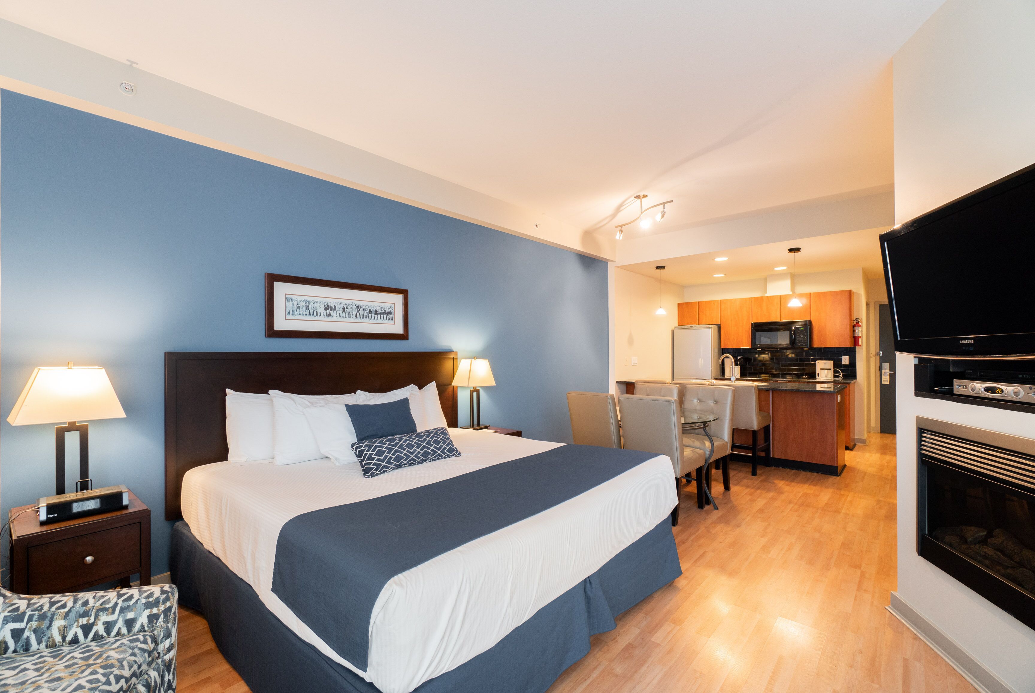 A hotel room with a large bed, a desk, a kitchenette, and a TV.